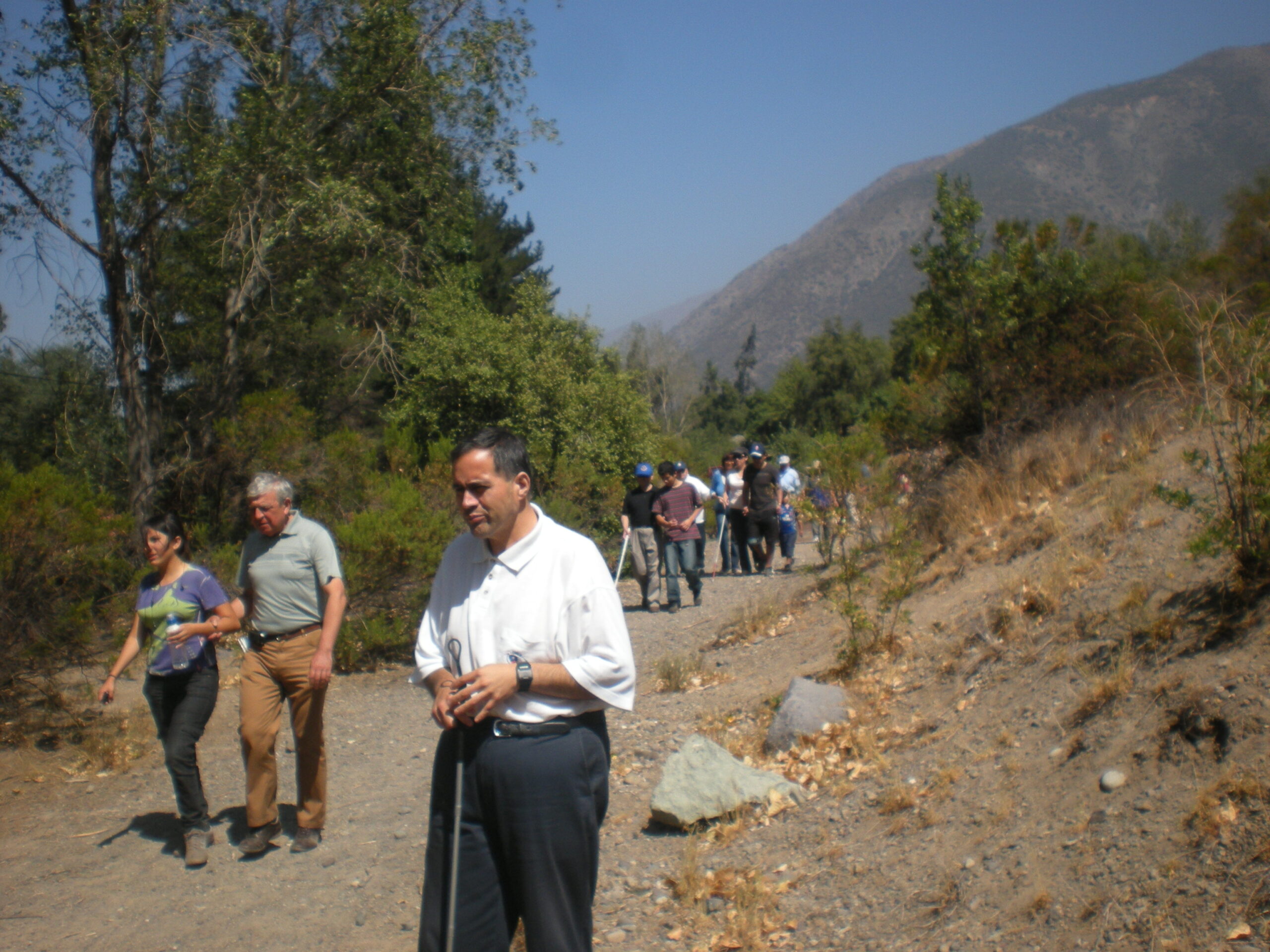 Personas ciegas acompañadas de guías caminan por un camino de tierra rodeado por árboles y piedras. De fondo se observa un cerro y el cielo despejado.