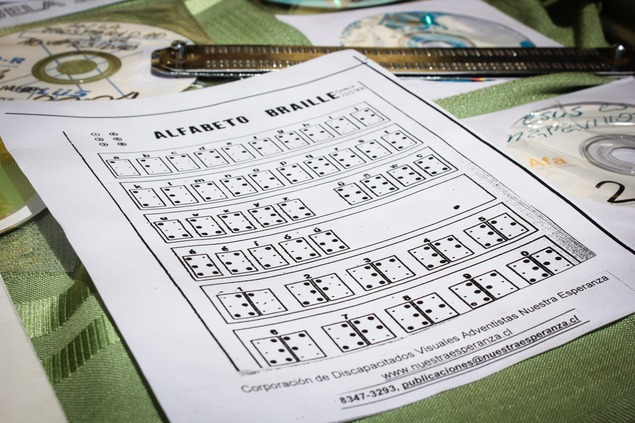 En una mesa, se observa de manera principal una hoja impresa, una regleta Braille y dos discos compactos con material en audio. La hoja de papel muestra gráficamente el alfabeto Braille.